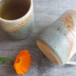 Może przedstawiać: Dwa ręcznie robione ceramiczne kubki o rustykalnym designie. Kubki mają kremową podstawę z niebieskimi i brązowymi akcentami. Jeden kubek jest w pozycji pionowej, wypełniony jasnym płynem, a drugi jest przechylony. Pomarańczowy kwiat jest na pierwszym planie.