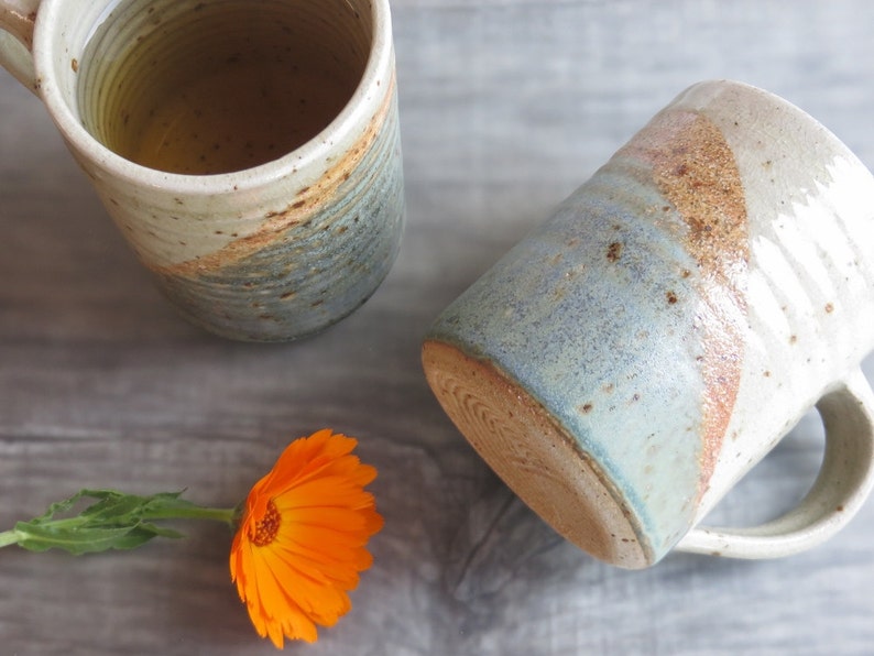 Może przedstawiać: Dwa ręcznie robione ceramiczne kubki o rustykalnym designie. Kubki mają kremową podstawę z niebieskimi i brązowymi akcentami. Jeden kubek jest w pozycji pionowej, wypełniony jasnym płynem, a drugi jest przechylony. Pomarańczowy kwiat jest na pierwszym planie.