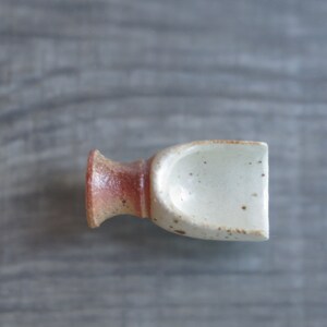 May include: A small, white ceramic scoop with brown speckles and a reddish-brown base. The scoop is designed for scooping small amounts of loose items, such as salt or sugar.