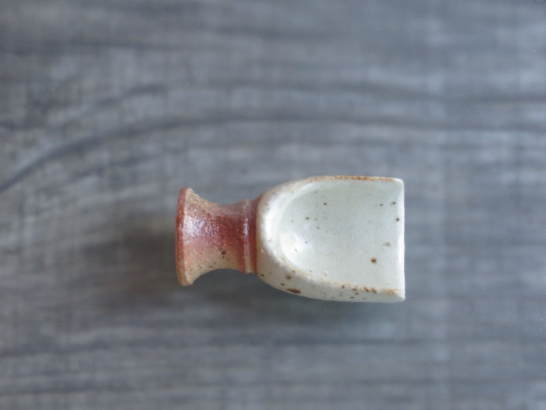 May include: A small, white ceramic scoop with brown speckles and a reddish-brown base. The scoop is designed for scooping small amounts of loose items, such as salt or sugar.
