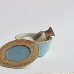 May include: A blue ceramic salt cellar with a lid and a small scoop. The cellar is filled with white salt.