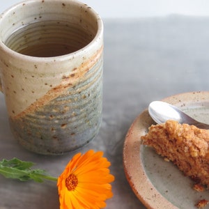 Może przedstawiać: Ręcznie robiony ceramiczny kubek z beżowo-szaro-niebieską glazurą, wypełniony herbatą. Kawałek deseru na talerzu z łyżeczką. Pomarańczowy kwiat dodaje koloru.