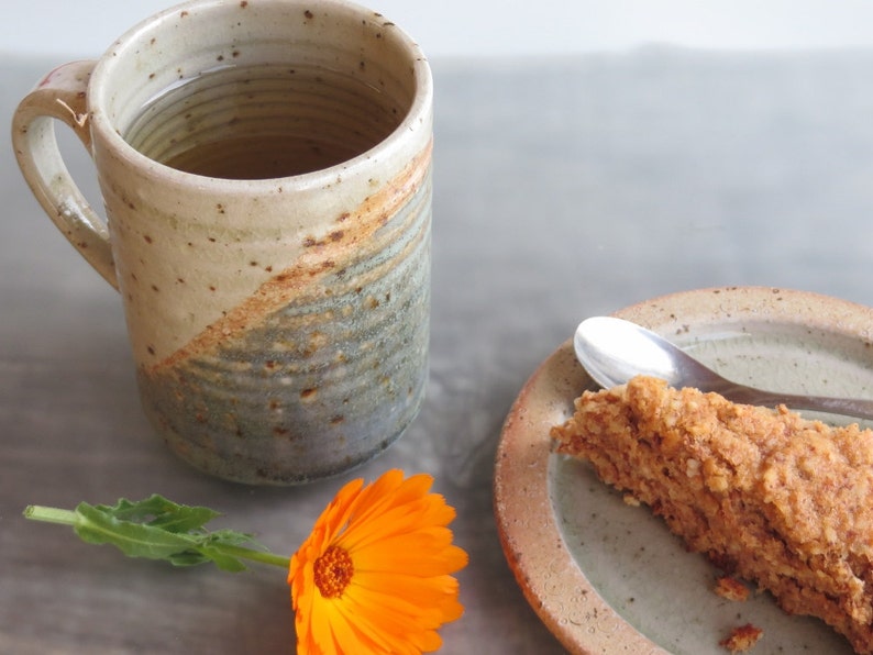 Może przedstawiać: Ręcznie robiony ceramiczny kubek z beżowo-szaro-niebieską glazurą, wypełniony herbatą. Kawałek deseru na talerzu z łyżeczką. Pomarańczowy kwiat dodaje koloru.