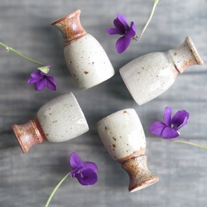 May include: Four small ceramic cups and vases with speckled white glaze and brown accents. The cups and vases are arranged on a wood surface with purple flowers.