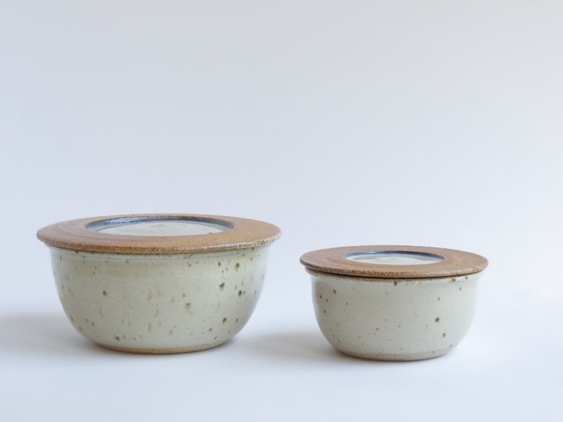 May include: Two ceramic butter dishes with lids. The larger dish is off-white with brown speckles and a brown lid. The smaller dish is the same color and has a brown lid.