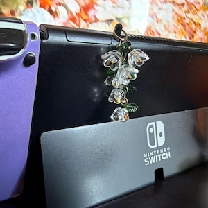 May include: A decorative charm featuring a cluster of translucent, clear glass-like flower blossoms with small green leaves, suspended from a black cord loop. The charm is attached to the top edge of a black Nintendo Switch console device.
