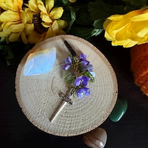 May include: A silver-toned sword charm adorned with small purple flower beads and green leaf accents, attached to a black cord. The charm is positioned next to a translucent, iridescent, angular stone on a circular, light-coloured wooden slice.