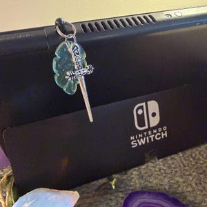 May include: A silver-toned metal sword charm attached to a small metal ring, featuring a translucent light green leaf-shaped pendant. The charm is hooked onto the top edge of a black Nintendo Switch console, which displays the white Nintendo Switch logo.