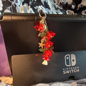 May include: A decorative charm featuring three translucent red flower-shaped beads, green leaf accents, a small gold-toned crescent moon, a gold-toned star, and a miniature red and white mushroom figurine, all suspended from a silver-toned metal ring.