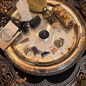 May include: A silver-toned metal tray containing a small straw broom with a bee-shaped handle, a clear quartz point, a black tourmaline stone, a sage bundle with yellow flowers, a black tea light candle, a glass vial, and several small plastic bags of herbs.