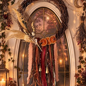 May include: A circular grapevine wreath adorned with dried pampas grass, white flowers, and three dried orange slices. Long, hanging fabric ribbons in shades of pink, red, brown, and green dangle from the base, accented with small gold metal bells.