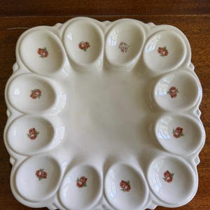 Könnte beinhalten: Eine quadratische, cremeweiße Eierservierplatte aus Keramik mit zwölf ovalen Vertiefungen, die in einem Rand angeordnet sind. Jede Vertiefung ist mit einem kleinen, mittigen Blumenmotiv aus roten Rosen und grünen Blättern verziert.