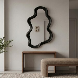 May include: A wall-mounted mirror featuring a thick, black, irregular wavy frame with an organic, fluid shape. The mirror is positioned above a rectangular wooden console table with clean lines and a smooth, natural wood grain finish.