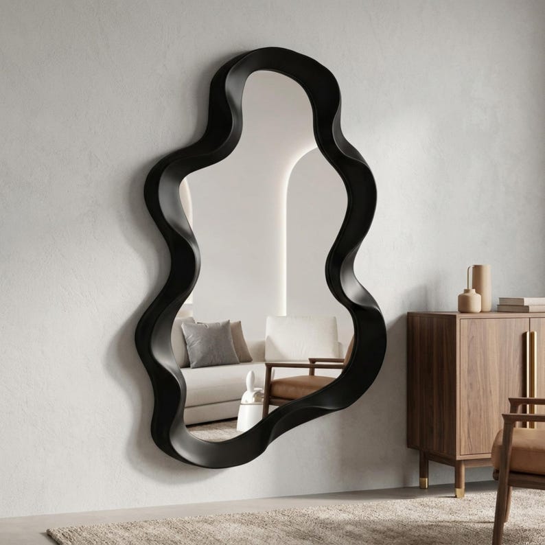 May include: A large wall mirror featuring a thick, matte black frame with an irregular, organic, wavy, or fluid shape. The reflective glass surface shows a partial view of a room interior, including a sofa, cushions, and a wooden cabinet.