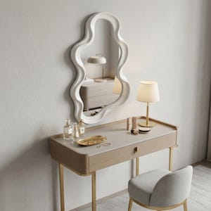 May include: A wall-mounted mirror with a white, wavy, organic-shaped frame positioned above a light wood vanity table. The vanity features a single drawer, gold-toned legs, a gold lamp, a gold tray with small items, and a matching gray upholstered chair.