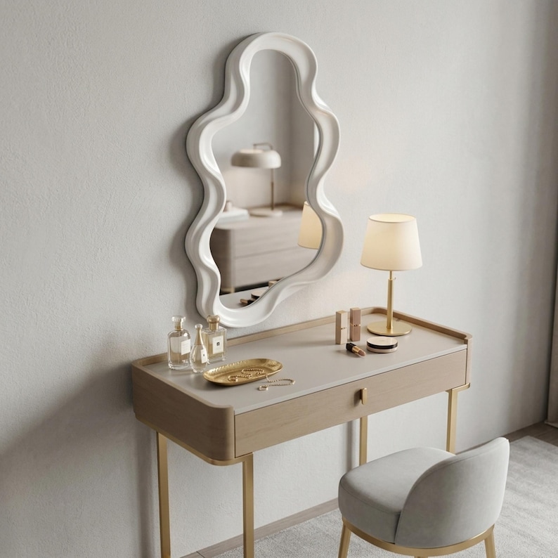 May include: A wall-mounted mirror with a white, wavy, organic-shaped frame positioned above a light wood vanity table. The vanity features a single drawer, gold-toned legs, a gold lamp, a gold tray with small items, and a matching gray upholstered chair.