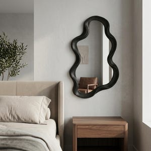 May include: A wall-mounted mirror featuring an irregular, wavy, organic-shaped frame in a matte black finish. The reflective glass surface is encased within the continuous, flowing border, creating a distinct silhouette against the wall.