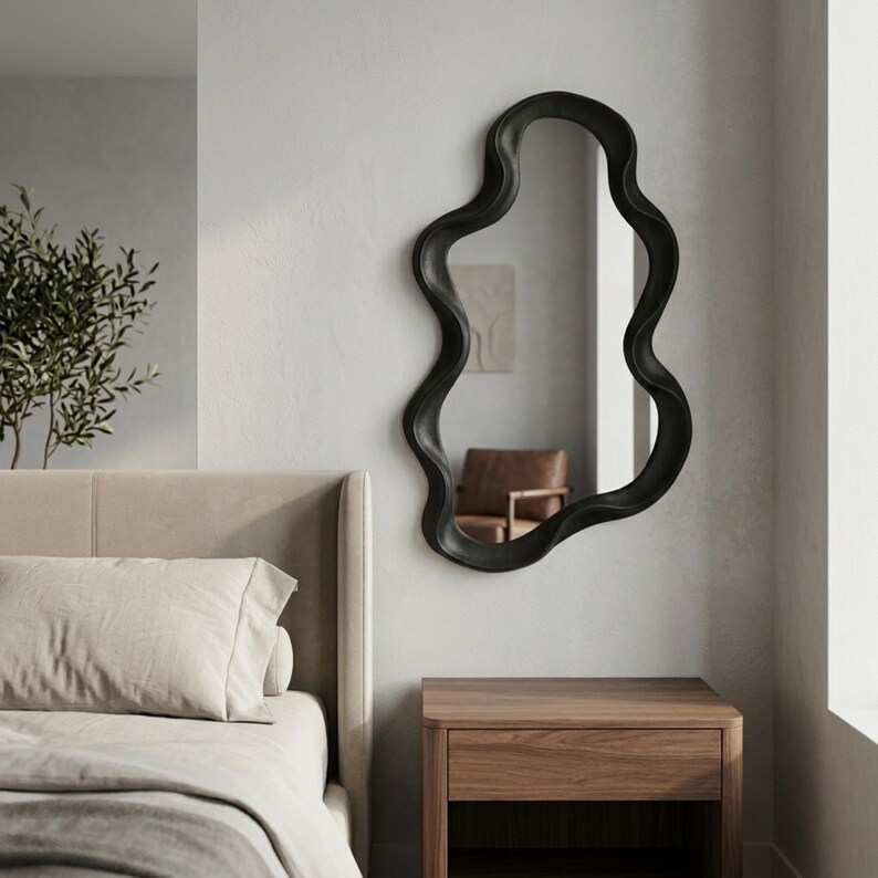 May include: A wall-mounted mirror featuring an irregular, wavy, organic-shaped frame in a matte black finish. The reflective glass surface is encased within the continuous, flowing border, creating a distinct silhouette against the wall.
