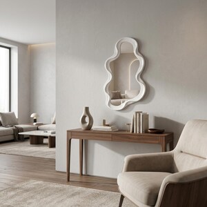 May include: A wall-mounted mirror with a white, irregular, wavy frame. The mirror has a fluid, organic shape and is positioned above a wooden console table. The reflective surface shows a room interior with neutral tones and soft lighting.