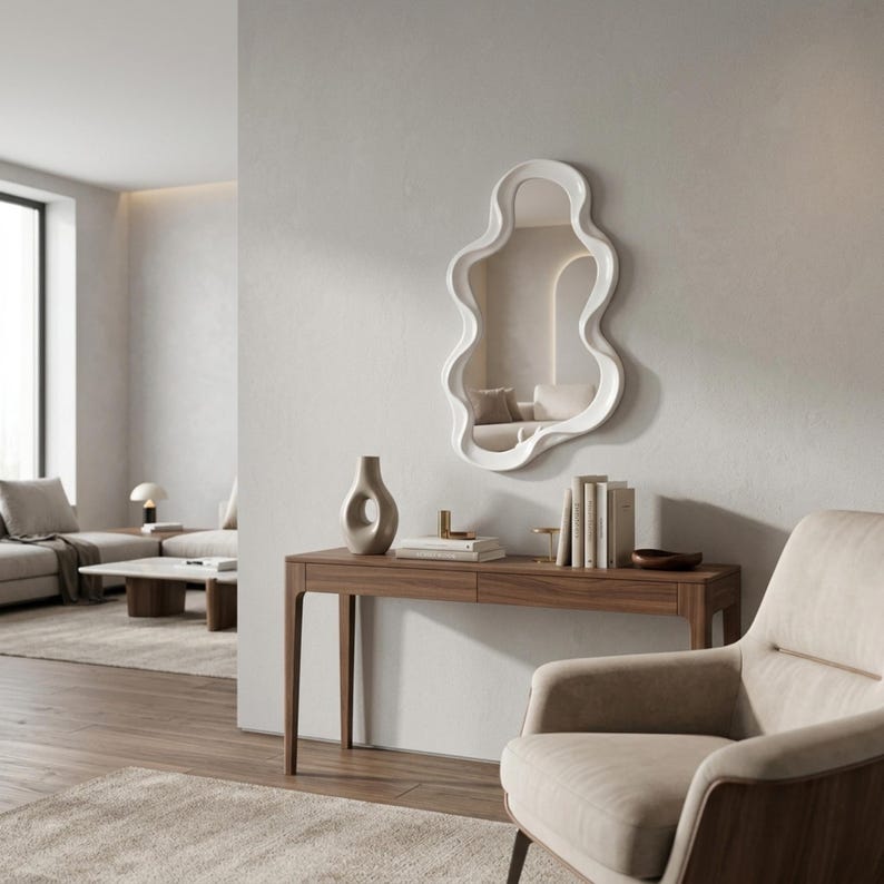 May include: A wall-mounted mirror with a white, irregular, wavy frame. The mirror has a fluid, organic shape and is positioned above a wooden console table. The reflective surface shows a room interior with neutral tones and soft lighting.