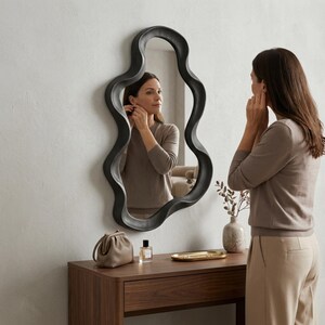 May include: A wall-mounted mirror with an irregular, wavy black frame. The reflective surface shows a reflection of a person wearing a beige long-sleeved top. The mirror is positioned above a wooden console table with a small bag, perfume, and tray.