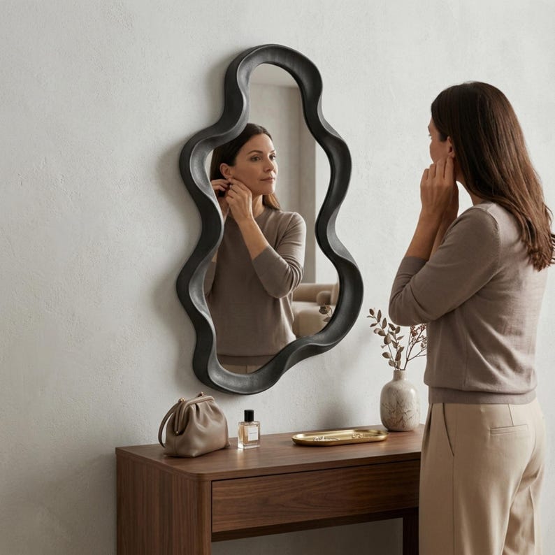 May include: A wall-mounted mirror with an irregular, wavy black frame. The reflective surface shows a reflection of a person wearing a beige long-sleeved top. The mirror is positioned above a wooden console table with a small bag, perfume, and tray.