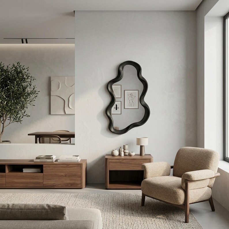 May include: An irregular, wavy-edged wall mirror with a thick black frame. The mirror is mounted on a light gray wall above a wooden side table. The reflection shows framed line art prints and a portion of a room with a light-colored wall.