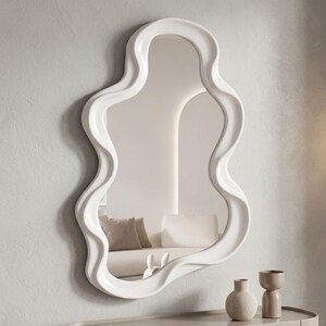 May include: A wall-mounted mirror featuring a thick, glossy white frame with an irregular, wavy, organic shape. The reflective glass surface shows a partial view of a room interior, including a sofa with cushions and a small decorative object.