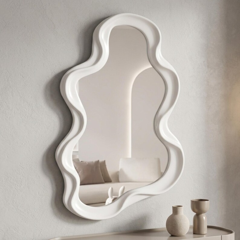 May include: A wall-mounted mirror featuring a thick, glossy white frame with an irregular, wavy, organic shape. The reflective glass surface shows a partial view of a room interior, including a sofa with cushions and a small decorative object.