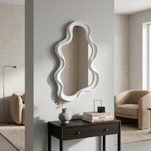 May include: A wall-mounted mirror featuring a thick, white, wavy, organic-shaped frame. The mirror is positioned above a dark brown wooden console table that holds a small gray vase and a stack of three books topped with a black and gold object.