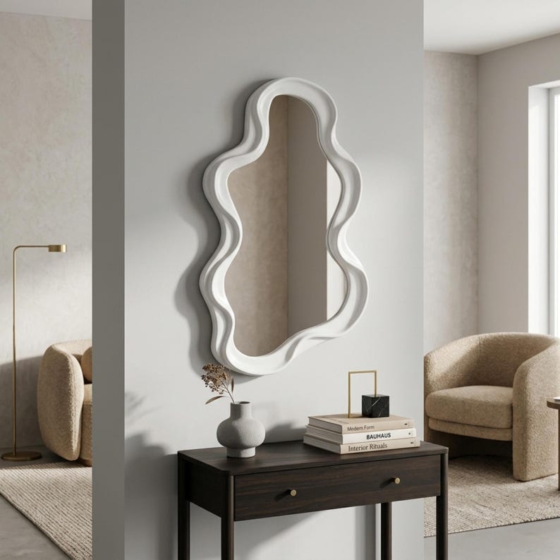 May include: A wall-mounted mirror featuring a thick, white, wavy, organic-shaped frame. The mirror is positioned above a dark brown wooden console table that holds a small gray vase and a stack of three books topped with a black and gold object.