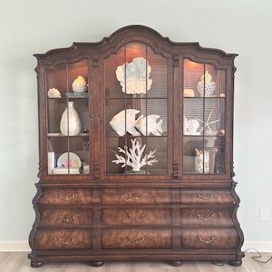 May include: A large, dark brown wooden china cabinet featuring a three-section glass display top with interior lighting and nine lower drawers with ornate brass-toned metal pulls. The shelves contain various white decorative coastal-themed items.