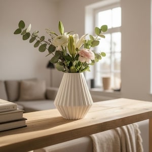 May include: A white ceramic vase with a geometric, vertically ribbed texture containing a floral arrangement of white lilies, pale pink ranunculus, and green eucalyptus branches, positioned on a light-colored wooden surface.