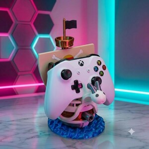 May include: A white Xbox controller rests on a stand shaped like a ship with a mast, a black flag, and a sail featuring a skull and crossbones emblem. The base is textured to resemble blue ocean waves, and a small white figure is attached to the front.