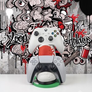 May include: A white and black gaming controller stand shaped like a sneaker, featuring a red heel tab and a green circular base. The stand holds a white Xbox controller on top and a white and black PlayStation controller on the bottom.