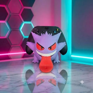 May include: A purple Gengar character figure serving as a controller stand, featuring red eyes, a wide white grin, and a long red tongue, positioned in front of a black gaming controller with colorful action buttons and a directional pad.