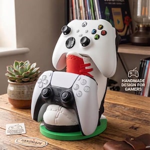 May include: A white and red sneaker-shaped controller stand holding a white Xbox controller on top and a white PlayStation controller in front. The base is green and circular. Text on the stand reads HANDMADE DESIGN FOR GAMERS.