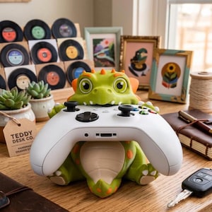 May include: A green dragon-shaped controller stand with orange spots and a cream-colored belly, holding a white wireless gaming controller. The dragon has large eyes, small horns, and is positioned to securely support the game controller.
