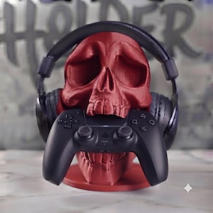 May include: A matte red skull-shaped stand designed to hold gaming accessories. The skull is wearing a pair of black over-ear headphones on its cranium, while a black wireless game controller is positioned in its mouth area for storage.
