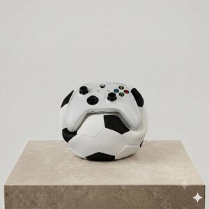 May include: A white wireless video game controller with black analog sticks and multicolored action buttons resting on top of a white and black soccer ball. The controller features the Xbox logo and standard directional inputs on its front surface.