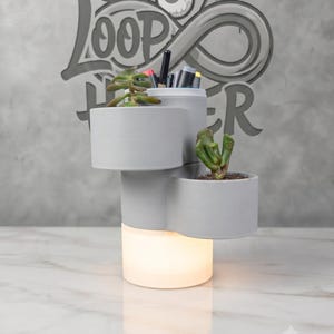 May include: A multi-tiered gray planter and desk organizer with a central illuminated base. The structure features three cylindrical compartments at varying heights, two containing small green succulent plants and one holding various pens and markers.