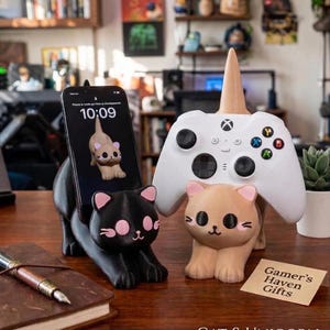 Op de afbeelding: Een smartphonehouder in de vorm van een zwarte kat en een controllerhouder in de vorm van een beige kat met een eenhoornhoorn. De zwarte kat houdt een smartphone met een kattenachtergrond vast, terwijl de beige kat een witte controller ondersteunt. Een kaartje toont Gamer's Haven Gifts.