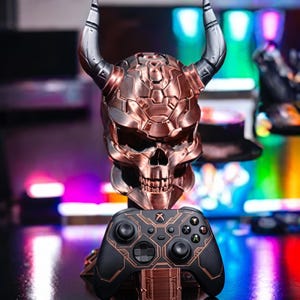 May include: A metallic copper-colored skull sculpture with large, dark grey horns, serving as a stand for a black wireless game controller. The controller features copper-colored geometric line patterns and standard buttons, including the Xbox logo.