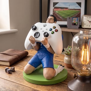以下が含まれることがあります： 青いユニフォームを着たサッカー選手のフィギュアが緑色の円形台座の上に膝をつき、黒いジョイスティックと色付きボタンが付いた白いワイヤレスゲームコントローラーを両手で顔の前に持っている様子。