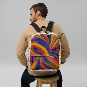Puede incluir: Una mochila con un vibrante patrón abstracto de remolinos en pinceladas azules, naranjas, amarillas y rojas. La bolsa tiene correas negras para los hombros, un bolsillo frontal con cremallera y un panel lateral crema con salpicaduras.