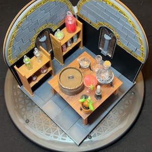 Może przedstawiać: Miniaturowa diorama pomieszczenia z kamiennymi ścianami, dwiema drewnianymi półkami z małymi szklanymi butelkami z kolorowymi płynami, centralnym drewnianym stołem z beczką, szklanymi kolbami, świecą i małą rośliną doniczkową na okrągłej podstawie.