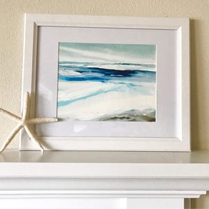May include: A framed watercolor painting of a seascape in shades of blue, white, and gray. The artwork is displayed in a white frame with a white mat. A white starfish rests on the shelf below the painting.