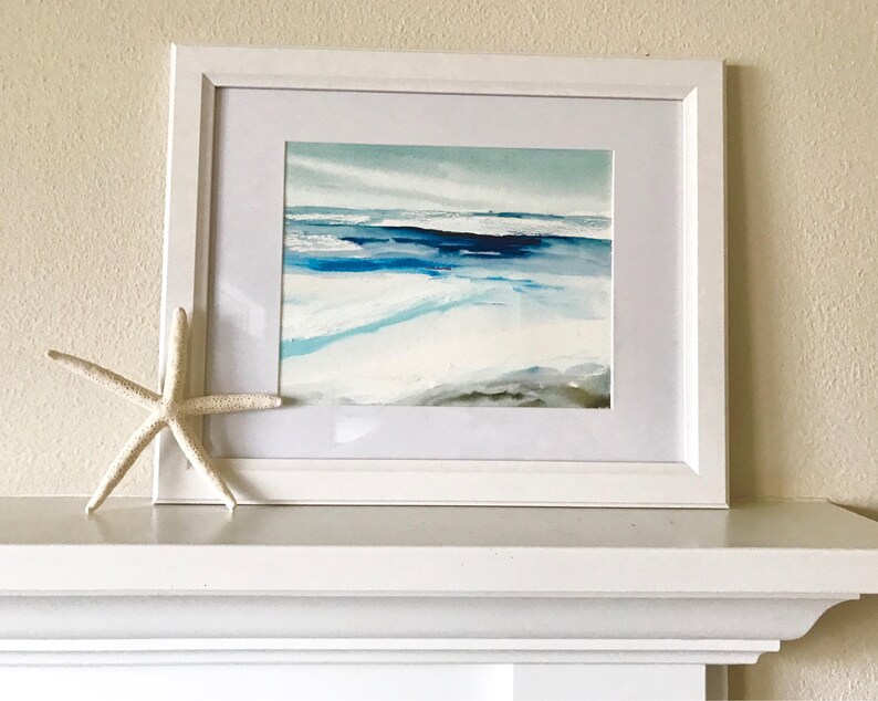 May include: A framed watercolor painting of a seascape in shades of blue, white, and gray. The artwork is displayed in a white frame with a white mat. A white starfish rests on the shelf below the painting.