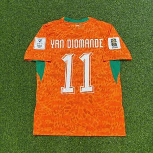 Pode incluir: Uma camisola de futebol laranja com um padrão animal subtil, o nome YAN DIOMANDE e o número 11 em branco. As mangas têm painéis laterais verdes, com um emblema FIFA Unite for Peace à esquerda e um emblema FIFA World Cup 2026 à direita.