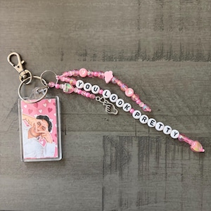 May include: A keychain featuring a rectangular clear acrylic charm with a photo of a person making a finger heart gesture against a pink heart background, attached to a silver lobster clasp and a strand of pink beads spelling YOU LOOK PRETTY.